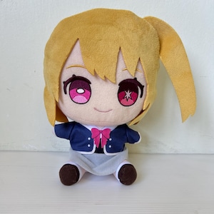 Może przedstawiać: Pluszowa lalka z jasnobrązowymi włosami i dużymi różowymi oczami. Nosi granatową kurtkę z różową kokardą, białą koszulę i szare szorty. Stopy lalki są brązowe.