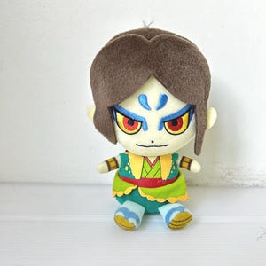Puede incluir: Muñeco de peluche con un personaje estilizado con cabello castaño, marcas faciales azules y ojos rojos. El muñeco viste un atuendo verde y amarillo con un cinturón rojo. El muñeco está sobre una superficie blanca.