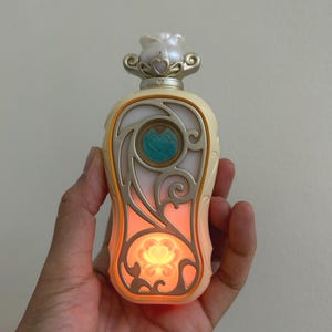 Puede incluir: Objeto decorativo iluminado con forma de botella, con cuerpo color crema y detalles dorados. El diseño presenta un motivo floral y un centro turquesa. La parte superior tiene un diseño de flor perlada. El interior brilla con luz naranja.
