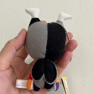 May include: A small plush doll with a black and gray body, white feet, and a silver chain. The doll has colorful accents on the bottom and a tag that reads "0147 MADE IN CHINA".