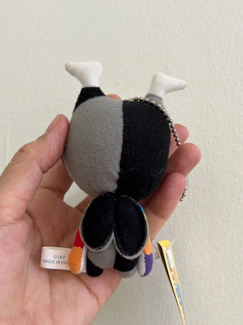 May include: A small plush doll with a black and gray body, white feet, and a silver chain. The doll has colorful accents on the bottom and a tag that reads "0147 MADE IN CHINA".