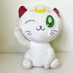 May include: A white plush cat with red ears, a yellow crescent moon on its forehead, and a winking green eye. The cat is in a seated position with one paw raised, and has black whiskers and a pink nose. A cute and cuddly plush toy.