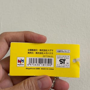 May include: A yellow tag with Japanese text and a barcode. The tag is attached to a silver ball chain. The text includes "MegaHouse" and "MADE IN CHINA". The tag also has a logo and the number "06".