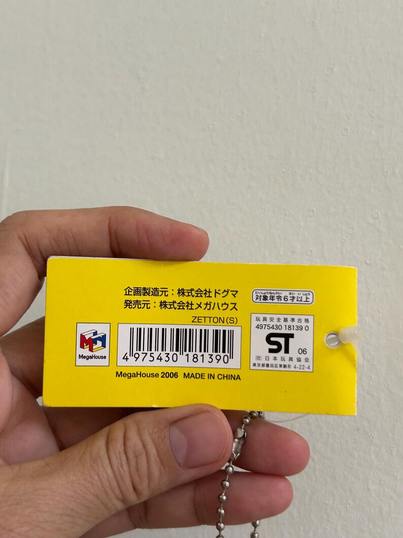 May include: A yellow tag with Japanese text and a barcode. The tag is attached to a silver ball chain. The text includes "MegaHouse" and "MADE IN CHINA". The tag also has a logo and the number "06".