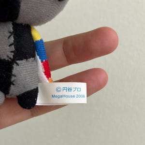 May include: A small, gray plush toy with black, patchwork details and colorful accents. The toy has a tag that reads "&copy; 円谷プロ MegaHouse 2006". The toy is held in a person's hand.