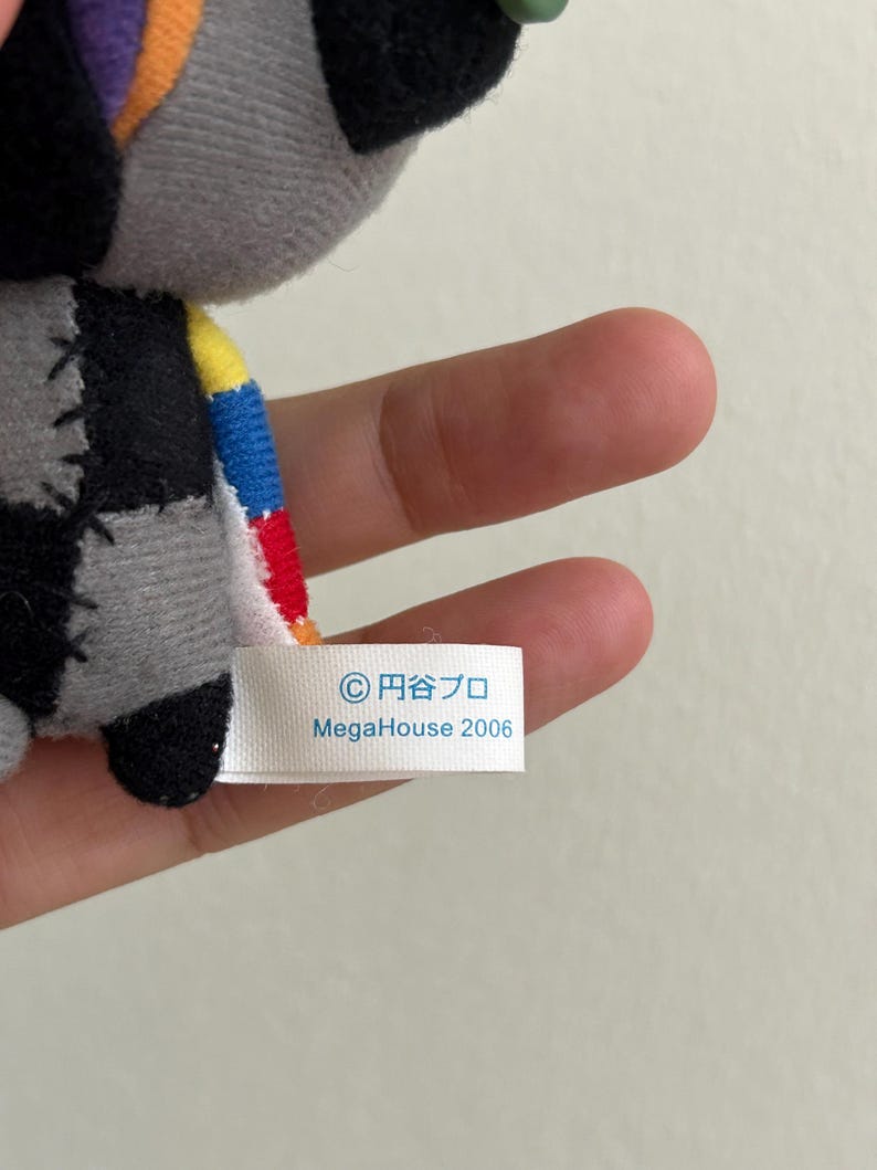 May include: A small, gray plush toy with black, patchwork details and colorful accents. The toy has a tag that reads "&copy; 円谷プロ MegaHouse 2006". The toy is held in a person's hand.