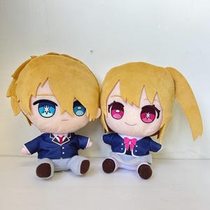 Op de afbeelding: Twee pluche poppen met anime-achtige kenmerken. De poppen hebben blond haar en grote ogen. De ene pop draagt een marineblauwe jas, een grijze broek en een rode stropdas. De andere pop draagt een marineblauwe jas, een roze strik en een grijze rok.