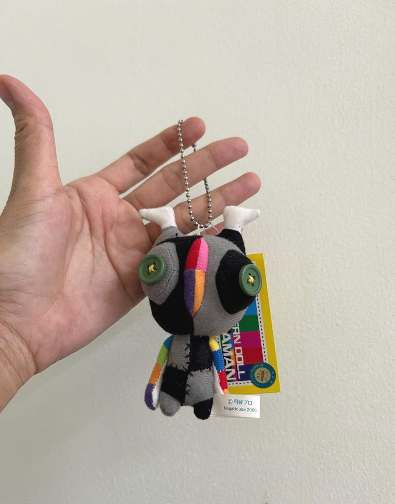May include: A small, handmade doll with a unique design. The doll has a gray body with black patches, button eyes, and a colorful striped head. It is attached to a silver chain and has a yellow tag with Japanese text.