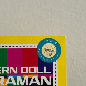 May include: A yellow box with a colorful striped design and the words "ERN DOLL RAMAN" in white. A blue circular sticker with Japanese text and a logo is affixed to the top right corner.