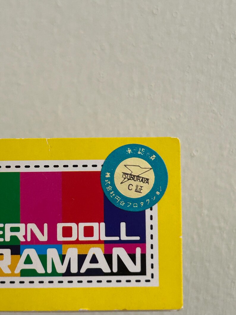 May include: A yellow box with a colorful striped design and the words "ERN DOLL RAMAN" in white. A blue circular sticker with Japanese text and a logo is affixed to the top right corner.