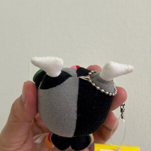May include: A small plush toy with a black and gray split design, white feet, and a silver chain. The toy has a colorful design on the bottom and a yellow tag with text. The plush is held in a person's hand.