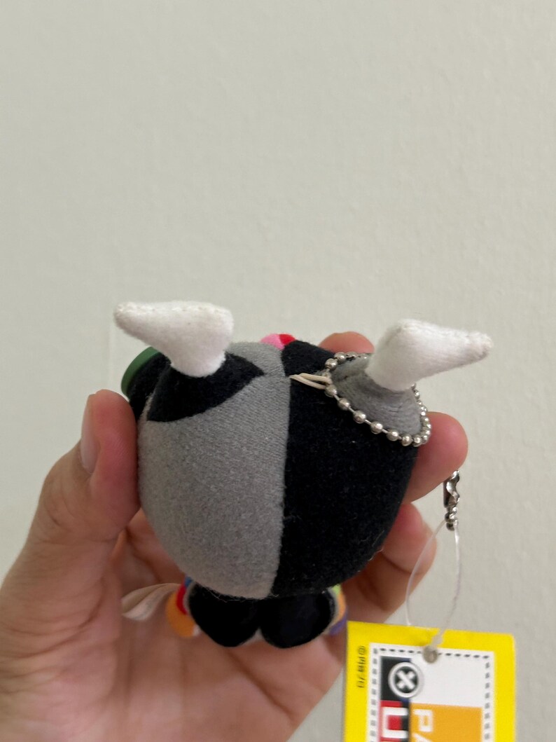 May include: A small plush toy with a black and gray split design, white feet, and a silver chain. The toy has a colorful design on the bottom and a yellow tag with text. The plush is held in a person's hand.