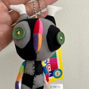 May include: A handmade doll with a patchwork design. The doll has a gray, black, and white body with colorful accents. It features green button eyes and a metal ball chain for hanging. A yellow tag is attached.