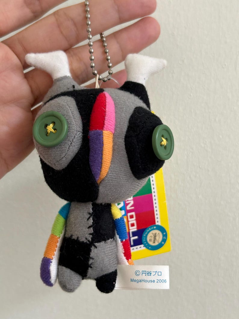 May include: A handmade doll with a patchwork design. The doll has a gray, black, and white body with colorful accents. It features green button eyes and a metal ball chain for hanging. A yellow tag is attached.