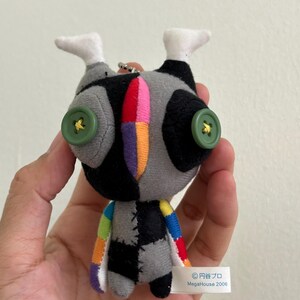 May include: A handmade plush doll with a patchwork design. The doll features a gray and black body, green button eyes, and colorful accents. The doll has white feet and a small tag that reads "MegaHouse 2006".