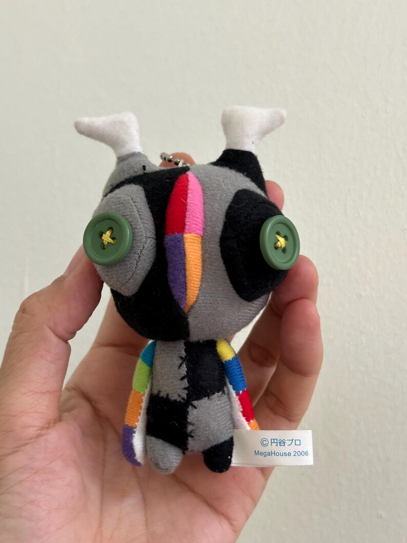 May include: A handmade plush doll with a patchwork design. The doll features a gray and black body, green button eyes, and colorful accents. The doll has white feet and a small tag that reads "MegaHouse 2006".