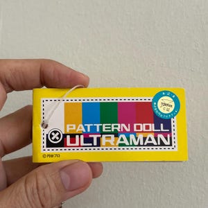 May include: A yellow rectangular tag with the text "PATTERN DOLL ULTRAMAN" in white. The tag has a color bar with yellow, blue, green, and pink sections. A small round sticker is in the upper right corner.