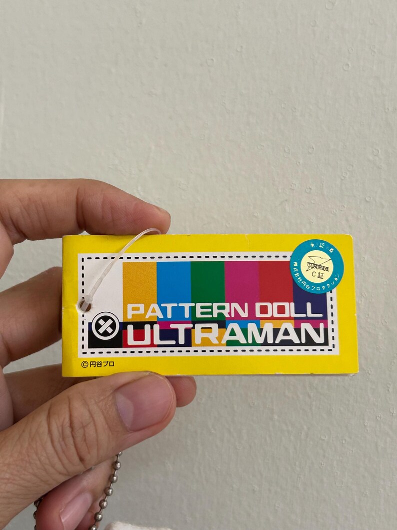 May include: A yellow rectangular tag with the text "PATTERN DOLL ULTRAMAN" in white. The tag has a color bar with yellow, blue, green, and pink sections. A small round sticker is in the upper right corner.
