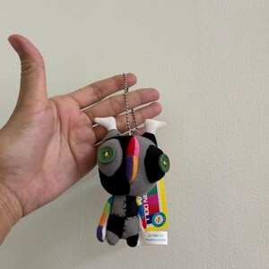 May include: A small, handmade doll with a patchwork design. The doll features a black head with large green button eyes, a gray body with black patches, and a rainbow-colored arm. It hangs from a silver chain with a tag.