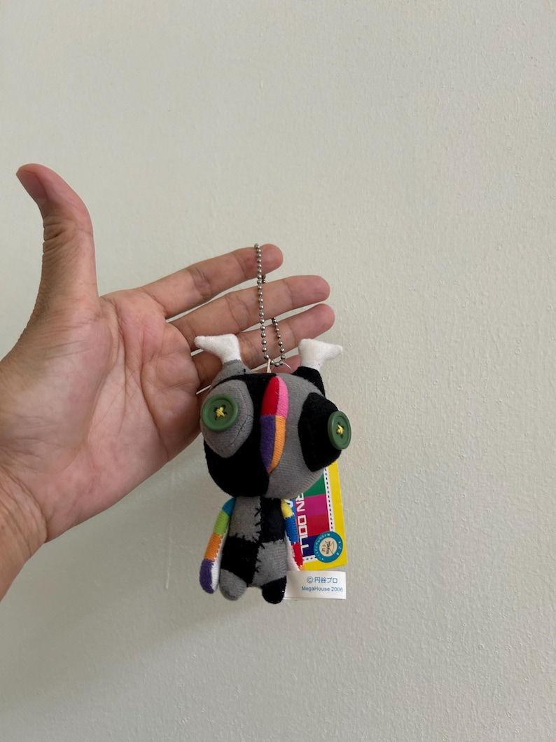 May include: A small, handmade doll with a patchwork design. The doll features a black head with large green button eyes, a gray body with black patches, and a rainbow-colored arm. It hangs from a silver chain with a tag.