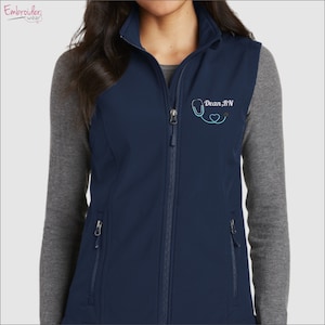 May include: A navy blue soft-shell vest with a zipper closure and two zippered pockets. The vest has a teal blue embroidered stethoscope with a heart and the text "Dean, RN".