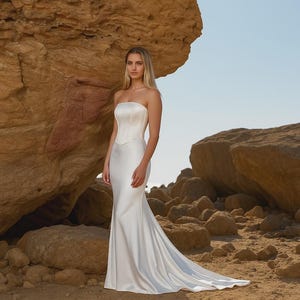 May include: A strapless, white satin wedding gown with a fitted bodice and a flowing mermaid skirt. The dress is worn by a person standing in a desert landscape with large rock formations and a clear blue sky.