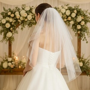 May include: A bride in a white strapless wedding dress and veil stands before a floral arch. The veil is sheer and falls to the shoulders. The background includes a wooden structure, candles, and floral arrangements.