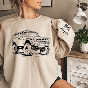 May include: A beige sweatshirt with a black graphic of a vintage off-road vehicle. The sweatshirt has a black American flag patch on the sleeve with the word "Angela" written on it.
