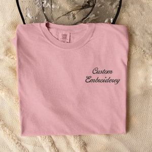 May include: A pale pink t-shirt with the text "Custom Embroidery" embroidered on the front in black.