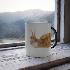 May include: White ceramic mug with a black handle featuring a watercolor illustration of a snail. The snail has a brown shell and a light pink body. Steam rises from the mug, suggesting a hot beverage.