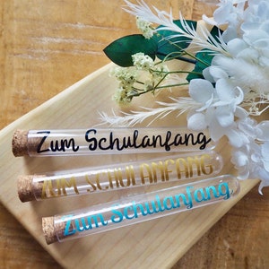 Op de afbeelding: Drie doorzichtige glazen reageerbuizen met kurkstoppen. De buizen zijn versierd met gouden en blauwe letters die "Zum Schulanfang" zeggen.