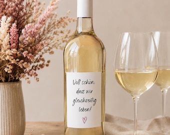 May include: A clear glass wine bottle with a white label featuring the black text "Voll schön, dass wir gleichzeitig leben!" and a small pink heart. The bottle is filled with a pale yellow liquid and has a white screw cap closure.