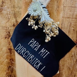 Può includere: Tessuto nero con testo bianco che recita "Papa mit Durchblick!" Il tessuto è su una superficie di legno con fiori bianchi secchi.