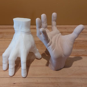 Puede incluir: Dos manos esculpidas, una blanca y otra color carne, se exhiben sobre una superficie de madera. La mano blanca está en una posición relajada, mientras que la mano color carne está abierta, como si se extendiera. Las manos son detalladas y parecen estar hechas de un material sólido.