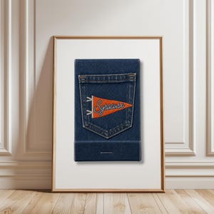 Puede incluir: Obra de arte enmarcada con un diseño de efecto denim y un bolsillo trasero. Un banderín naranja con la palabra "Syracuse" en escritura blanca está en el bolsillo. La obra está en un marco blanco contra una pared blanca y un suelo de madera.