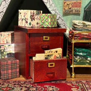 Pu&ograve; includere: Una cassettiera in miniatura in legno rosso scuro a due cassetti con maniglie in metallo dorato. Il cassetto superiore &egrave; chiuso e quello inferiore &egrave; socchiuso, rivelando un piccolo libro intitolato Life. Nelle vicinanze si trovano scatole regalo in miniatura e un carrello in metallo con tessuti piegati.