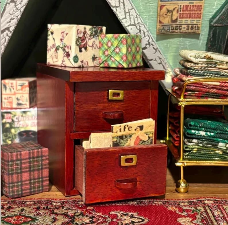Pu&ograve; includere: Una cassettiera in miniatura in legno rosso scuro a due cassetti con maniglie in metallo dorato. Il cassetto superiore &egrave; chiuso e quello inferiore &egrave; socchiuso, rivelando un piccolo libro intitolato Life. Nelle vicinanze si trovano scatole regalo in miniatura e un carrello in metallo con tessuti piegati.