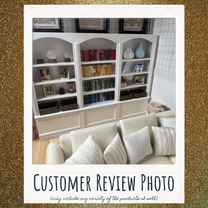 May include: A large white wooden bookshelf with three sections filled with books arranged by color, decorative vases, framed photographs, and woven baskets. In front of the bookshelf sits a cream-colored sofa with several textured throw pillows.