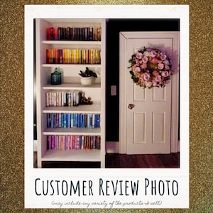 May include: A white bookshelf filled with rows of books organized by color, featuring a small potted plant and a glass jar on the shelves. Next to the bookshelf is a white door with a decorative floral wreath made of pink and white flowers.