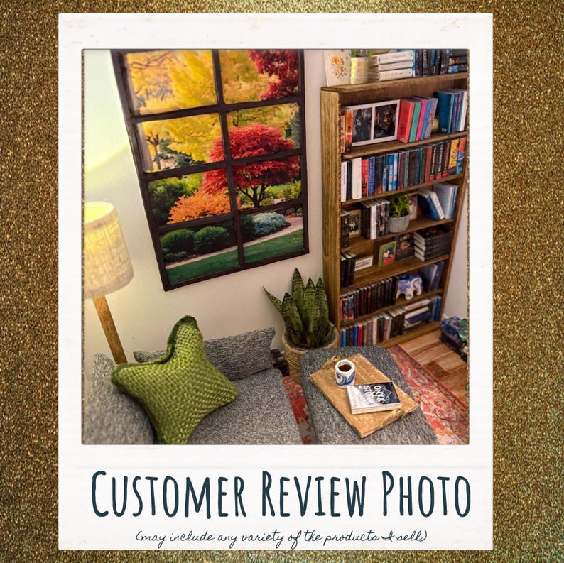 May include: A room interior featuring a wall-mounted window-style art print displaying an autumn landscape, a tall wooden bookshelf filled with books, a gray upholstered armchair with a green textured pillow, and a wooden side table with a book.