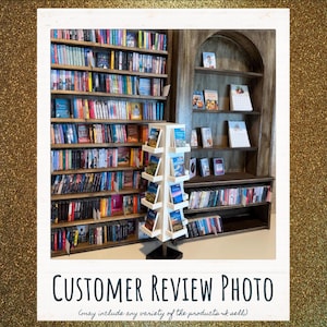 May include: A tall, white, four-sided rotating display rack filled with books stands in front of a large, dark wood bookshelf. The bookshelf features multiple horizontal shelves packed with various books, while an arched wooden unit displays select titles.