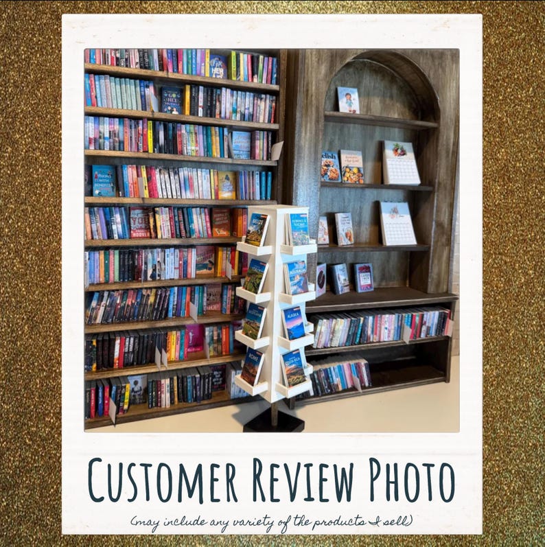 May include: A tall, white, four-sided rotating display rack filled with books stands in front of a large, dark wood bookshelf. The bookshelf features multiple horizontal shelves packed with various books, while an arched wooden unit displays select titles.