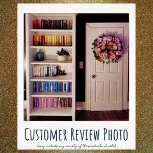 May include: A white bookshelf with five shelves filled with books organized by color, a small teal glass jar, a potted plant, and a decorative object. Next to the bookshelf is a white door with a floral wreath featuring pink and white flowers.