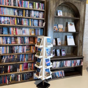 May include: A tall, white, four-sided rotating floor display stand filled with travel guidebooks, positioned in front of large, dark wood bookshelves packed with numerous books arranged on multiple horizontal shelves in a bookstore setting.