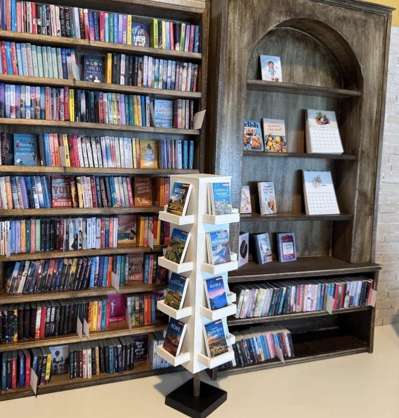 May include: A tall, white, four-sided rotating floor display stand filled with travel guidebooks, positioned in front of large, dark wood bookshelves packed with numerous books arranged on multiple horizontal shelves in a bookstore setting.