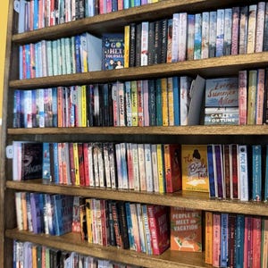 May include: A wooden bookshelf filled with numerous books arranged in rows. The spines display various colors, titles, and authors. Some books are positioned facing forward, including titles like Go Hex Yourself, Every Summer After, The Bride Test, and People We Meet on Vacation. Alphabetical dividers are visible on the shelves.