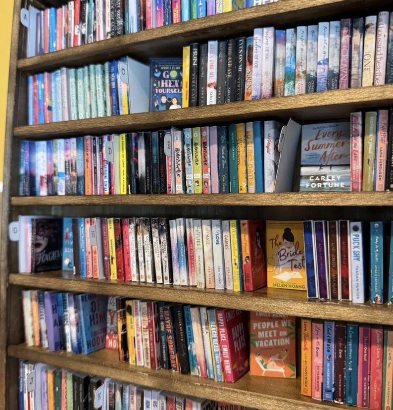 May include: A wooden bookshelf filled with numerous books arranged in rows. The spines display various colors, titles, and authors. Some books are positioned facing forward, including titles like Go Hex Yourself, Every Summer After, The Bride Test, and People We Meet on Vacation. Alphabetical dividers are visible on the shelves.