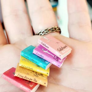 May include: A stack of six miniature books with colorful covers including pink, purple, teal, yellow, tan, and red. Each book features printed titles and author names on the front covers, including Solitaire, Loveless, Radio Silence, and I Was Born for This.