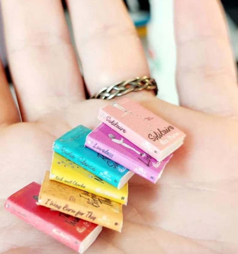 May include: A stack of six miniature books with colorful covers including pink, purple, teal, yellow, tan, and red. Each book features printed titles and author names on the front covers, including Solitaire, Loveless, Radio Silence, and I Was Born for This.