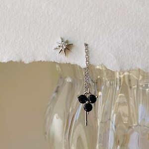 May include: A pair of silver earrings. One earring features a star with a single clear stone in the center. The other earring features a dangling chain with three black stones and a key charm.